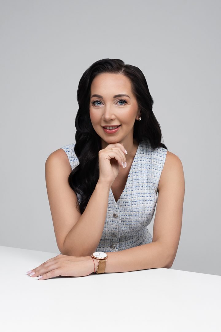 Asteri įkūrėja Joana Šaulienė, vilkinti šviesiai mėlyną liemenę, sėdi prie balto stalo.
